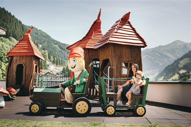A playground with colorful playhouses and a small train attraction. A cheerful child is sitting next to a mascot and enjoying time outdoors.