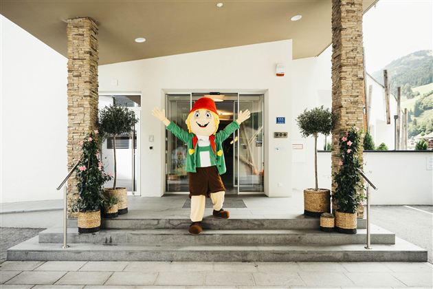 A cheerful mascot figure stands in front of a building and greets visitors. Surrounded by plants and an inviting atmosphere, it appears friendly and welcoming.