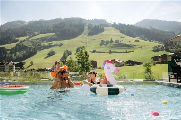 A family is having fun in the pool with an airy unicorn swing. In the background, green hills and traditional houses can be seen.