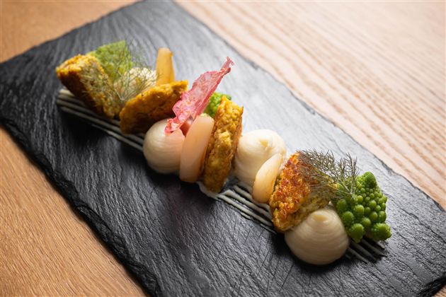 An artistically arranged dish on a black stone. It contains fried pieces, creamy side dishes, and fresh vegetables.