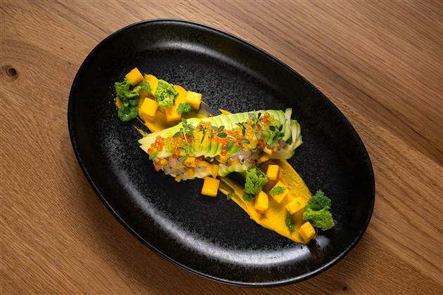 An artistic dish on a black plate. It consists of sushi, broccoli, and fresh mango.