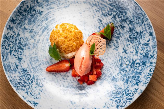 An artfully arranged dessert on a blue plate. It contains a yellow cream, strawberries, and a pink sorbet.