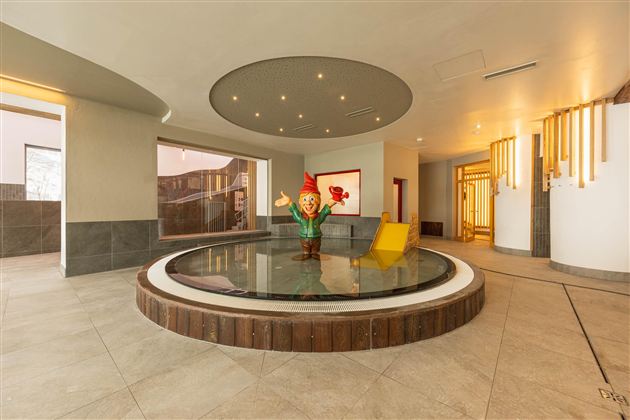A modern reception area with a round water basin in the center. In the middle stands a colorful figure with a red hat and a yellow lounge chair.