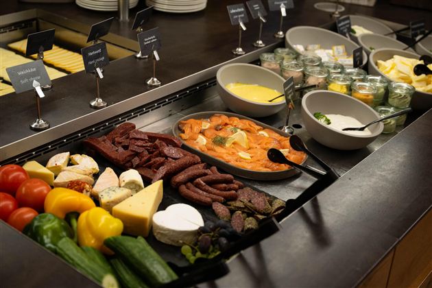 A rich buffet with various types of meat, cheese, fresh vegetables, and dips. Next to it, there are several bowls of desserts and a creamy sauce.