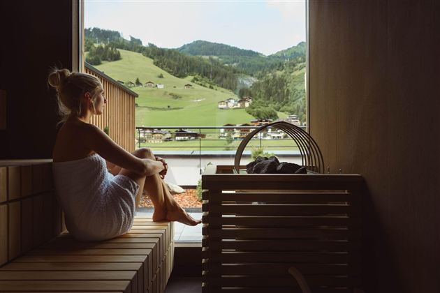 A woman is sitting relaxed in a sauna and enjoying the view of green hills. The atmosphere is calm and inviting.