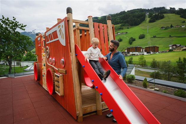 A child happily slides down a slide of a colorful play structure. In the background, green hills and a beautiful landscape can be seen.