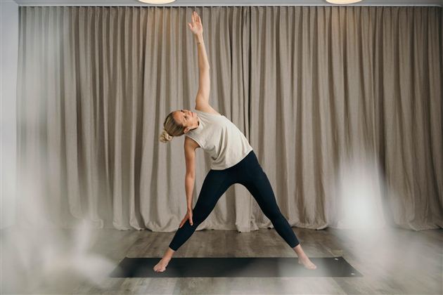 A woman is performing a yoga exercise in a bright room. The room has a neutral background with curtains.