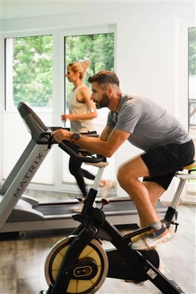 A man is training on a stationary bike while a woman jogs on a treadmill in the background. The room has large windows that let in natural light.