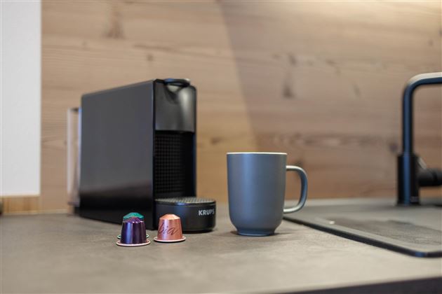 Eine moderne Kaffeemaschine steht auf einem Küchenarbeitsplatz. Daneben befinden sich zwei Kaffeekapseln und eine graue Tasse.