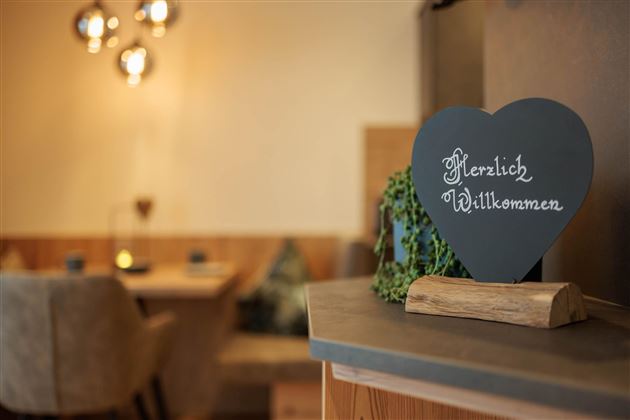 Ein herzförmiges Willkommensschild steht auf einem Tisch. Im Hintergrund sind ein gemütlicher Sitzbereich und sanfte Lichtquellen zu sehen.