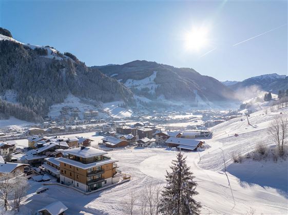 Eine schneebedeckte Berglandschaft mit einem kleinen Dorf und vielen Häusern. Die Sonne scheint über die Berge und die Skigebiete sind sichtbar.