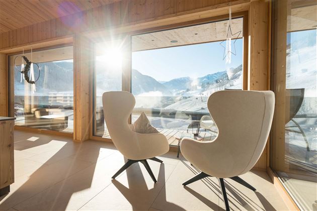 Helle, moderne Sitzecke mit zwei bequemen Stühlen und großen Fenstern, die einen Blick auf die Berge bieten. Die Sonne strahlt herein und sorgt für eine warme, einladende Atmosphäre.