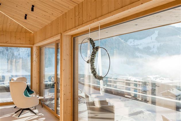 Ein modernes, lichtdurchflutetes Zimmer mit großen Fenstern und Holzverkleidung. Im Hintergrund sind verschneite Berge und ein winterlicher Ausblick zu sehen.