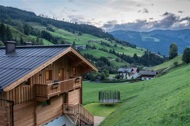 Ein schönes Holzhaus in einer grünen Hügellandschaft. Im Hintergrund sind Berge und kleine Dörfer zu sehen.