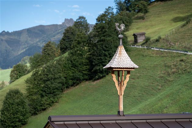 Ein traditioneller Dachreiter mit einer Wetterfahne, der auf einem modernen Gebäude steht. Im Hintergrund sind sanfte Hügel und Bäume vor einer Bergkulisse zu sehen.