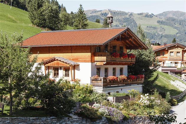 A charming chalet in the Alps with a red roof and blooming flowers. Surrounded by green meadows and wooded mountains.