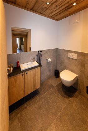 A modern bathroom with a wooden ceiling and elegant tiles. It features a sink, a toilet, and a large mirror.