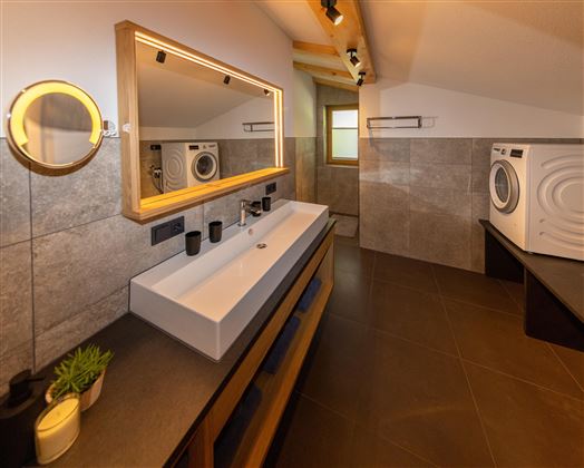 A modern bathroom with a large sink and a mirror surrounded by soft light. On the right side, there is a washing machine.
