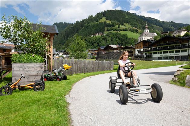 Ein Mädchen fährt in einem Kettcar auf einer Straße in der Natur. Im Hintergrund sind Berge und typische alpine Häuser zu sehen.