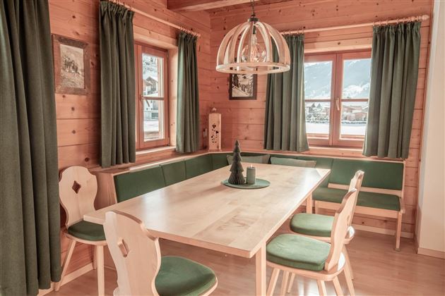 A cozy dining area with a wooden table and chairs in a rustic setting. Large windows let in plenty of light and create a warm atmosphere.