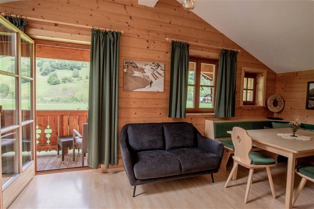 Ein gemütliches Zimmer in einem Holzhaus mit Blick auf die grüne Landschaft. Es gibt ein Sofa und einen Esstisch mit Stühlen.