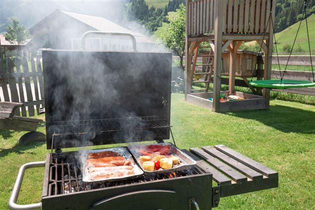 Ein Grill steht im Grünen und raucht, während Würstchen und Gemüse darauf garen. Im Hintergrund sieht man einen Spielplatz und eine malerische Landschaft.