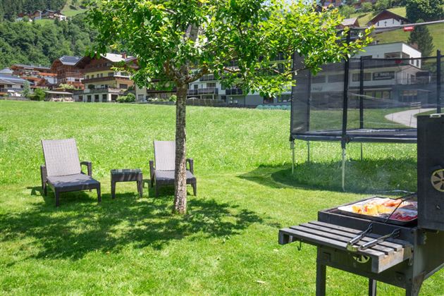 Ein ruhiger Garten mit zwei Liegestühlen, einem Grill und einem Trampolin. Im Hintergrund sind Häuser und grüne Berge zu sehen.