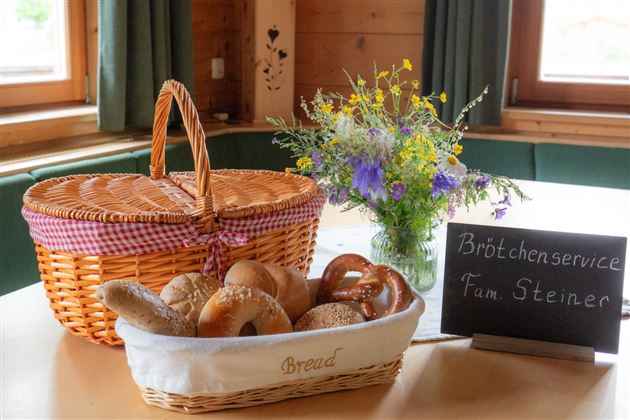 Ein Korb mit frischen Brötchen und Brezeln steht auf einem Tisch. Daneben steht ein Blumenstrauß und eine Tafel mit dem Hinweis "Brötchenservice Fam. Steiner".