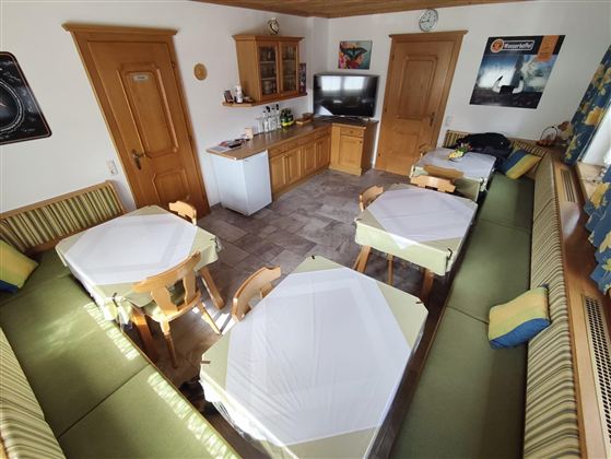 Ein gemütlicher Speisebereich mit mehreren Tischen und Sitzbänken. Die Einrichtung ist hell und freundlich, mit einer modernen Küche im Hintergrund.