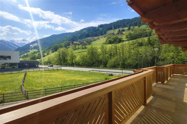 Eine schöne Aussicht von einem Balkon auf grüne Wiesen und Berge. Die Sonne scheint und der Himmel ist blau mit wenigen Wolken.