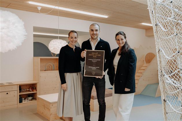 Three people are standing in a bright room holding an award. In the background, there are play equipment and wooden shelves.