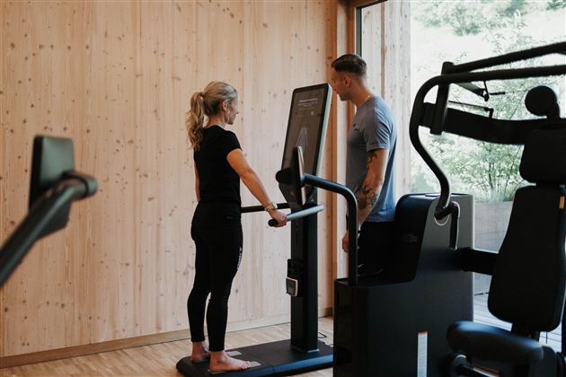 A woman stands at a fitness machine while a man gives her instructions. The room has wooden walls and large windows that let in a lot of light.