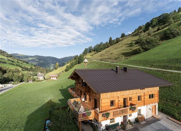 Ein Holzhaus umgeben von grünen Wiesen und sanften Hügeln. Der Himmel ist klar mit wenigen Wolken.