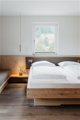 A modern bedroom with a wooden bed and a cozy seating area. The window offers a view outside to the landscape.