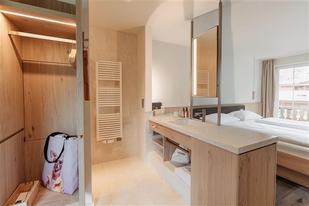 A modern bathroom with a bright wood cabinet and a large mirror. In the background, a cozy bedroom with a large bed is visible.