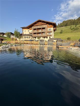 A beautiful hotel by the water, surrounded by mountains and green grass. The sky is blue and the atmosphere is calm.