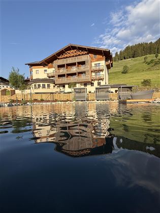 A beautiful hotel by the water with a wood-paneled facade. In the background, lush green hills and a blue sky can be seen.