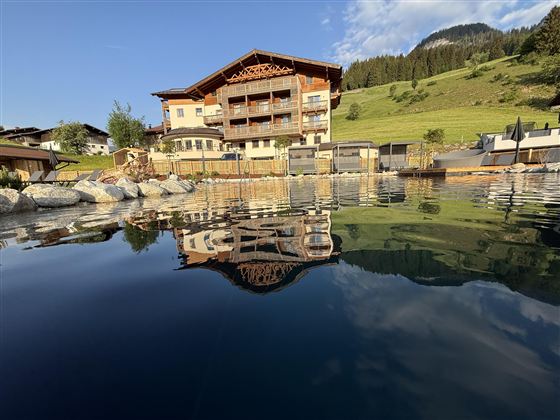 A beautiful hotel with a wooden facade is located by the water. The surrounding landscape is green and mountainous.