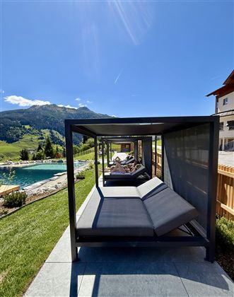 A quiet outdoor area with loungers and a pool. The surrounding landscape is green and mountainous under a clear blue sky.