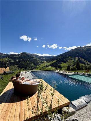 A relaxing panorama with a pool and a wooden terrace. In the background, green mountains and a clear blue sky can be seen.