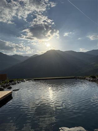 A tranquil pool extending before majestic mountains, under a clear sky with radiant sun. The gentle waves of the water reflect the surrounding landscape.