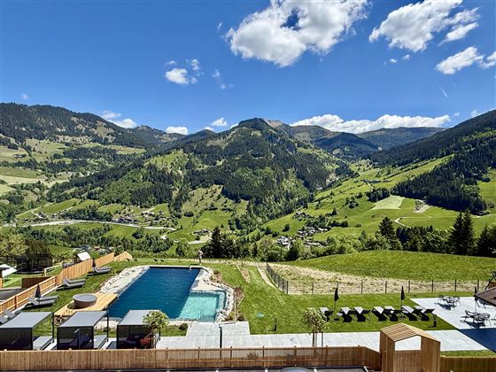 A beautiful landscape with green hills and mountains. In the foreground, there is a pool surrounded by loungers.