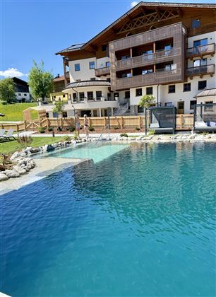 A beautiful outdoor pool with clear blue water and a wooden terrace. In the background, a modern building with balconies and a garden can be seen.