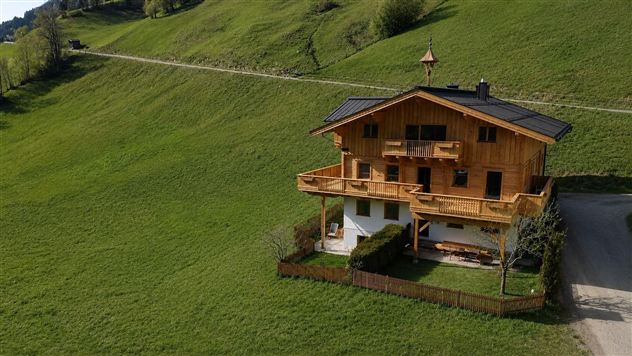 Ein schönes Holzhaus auf einer grünen Wiese. Die Umgebung ist von sanften Hügeln und einem klaren Himmel umgeben.