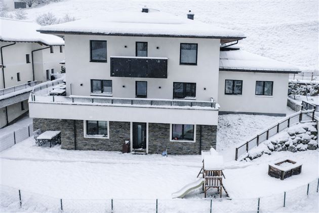 A modern house in the snow with a large balcony and a playground in the foreground. The surroundings are quiet and wintry.