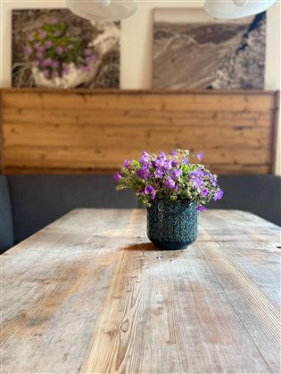 Ein Holztisch mit einer Vase voller lila Blumen steht im Vordergrund. Im Hintergrund sind Holzpaneele und Fotos an der Wand zu sehen.