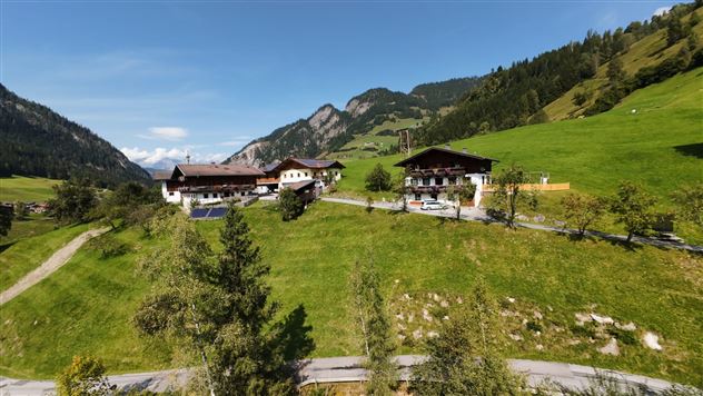 Eine malerische Landschaft mit grünen Wiesen und sanften Hügeln. Im Hintergrund sind Berge zu sehen, und einige traditionelle Gebäude stehen verstreut.