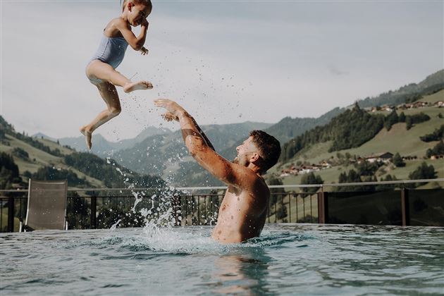 A father lifts his child into the air in the pool. In the background, green hills and a serene landscape can be seen.