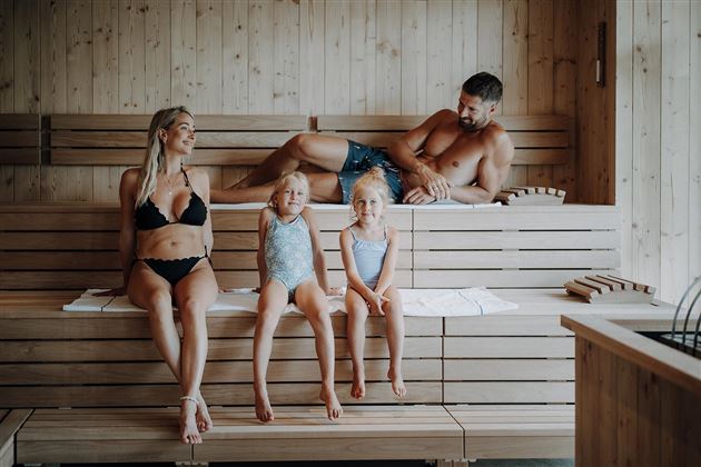 A family is relaxing in a sauna. Two girls and their parents are sitting on the wooden benches and enjoying their time together.