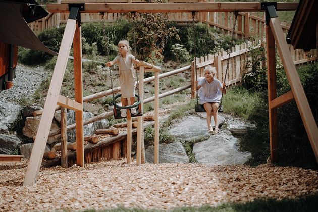 Two children are playing outdoors in a playground with a swing and a seesaw. The surroundings are green and designed to be close to nature.